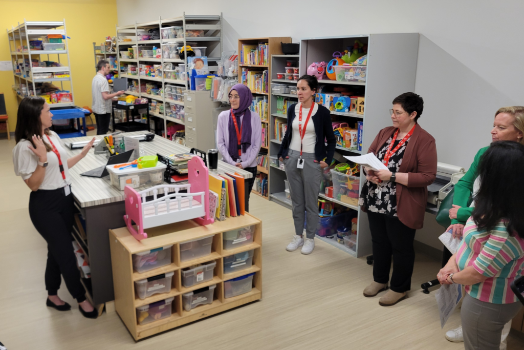 A room of bookcases filled with toys and individuals listening to another individual on a tour of KidsAbility's staff Resource Room. 