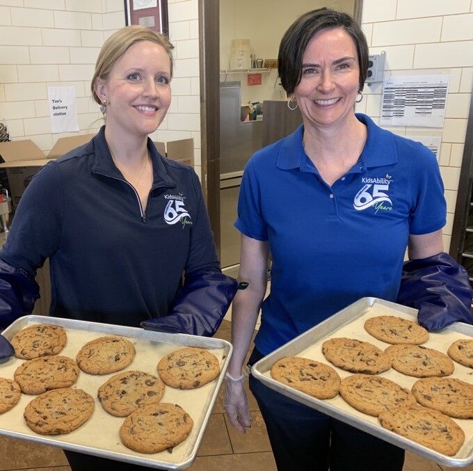 two women hold trays of chocolate chip cookies decorated with smiles