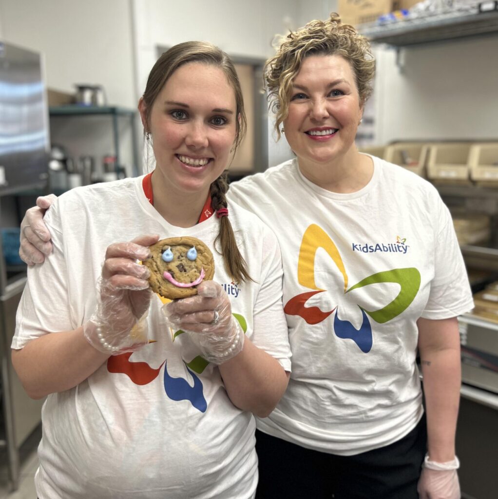 two woman wear volunteer t-shirts and hold a cookie with a decorated smile.